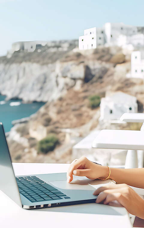 Frau sitzt mit ihrem Laptop draußen auf einer Terrasse, im Hintergrund sieht man das Meer 