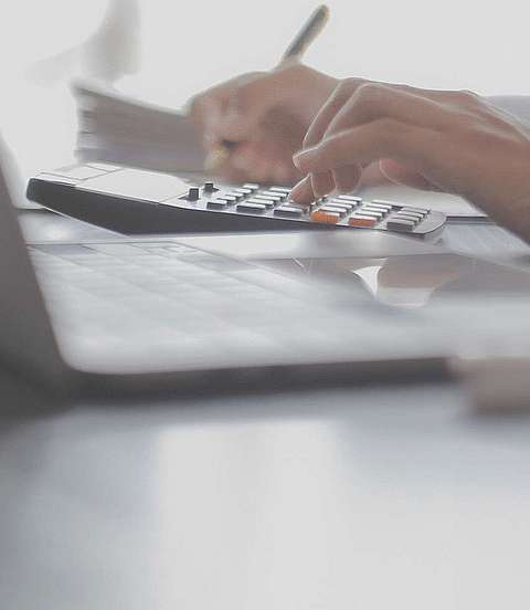 Frau sitzt am Schreibtisch, notiert sich etwas und tippt auf einen Taschenrechner