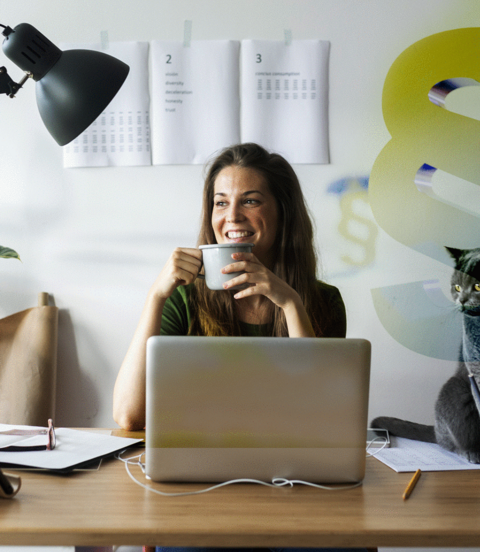 Frau sitzt am Schreibtisch, hält eine Tasse in der Hand und lächelt